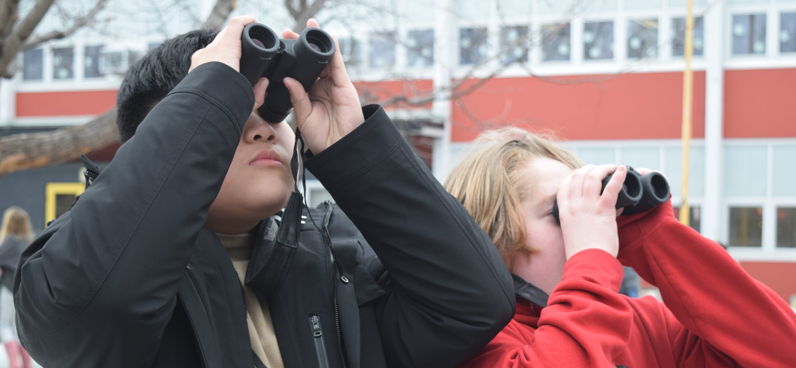 Two students outside their school holding binoculars up to their eyes.